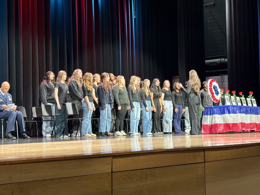 A group of mostly young women in formation stand in a line or semicircle on a stage while a woman (conductor/speaker) faces them. To the right is a table draped in red, white, and blue with framed photos, flowers, and a large wreath—indicating a Veterans' Day memorial display. On the far left a man in a JROTC uniform sits reading from a paper; the background is a black curtain.
