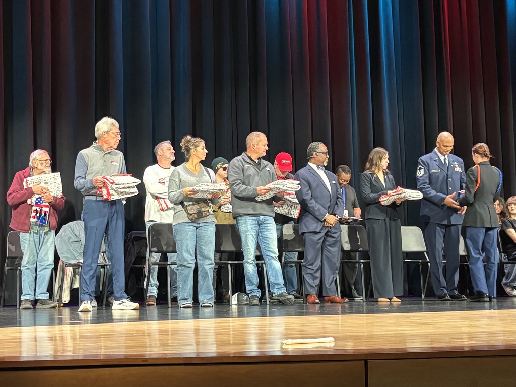A group of people stand on a stage in front of a black curtain holding quilts. Chairs sit behind them on a wooden floor. Attire ranges from casual to formal; one person in a JROTC uniform is shaking hands with a woman. The scene appears to be part of a Veterans' Day ceremony.