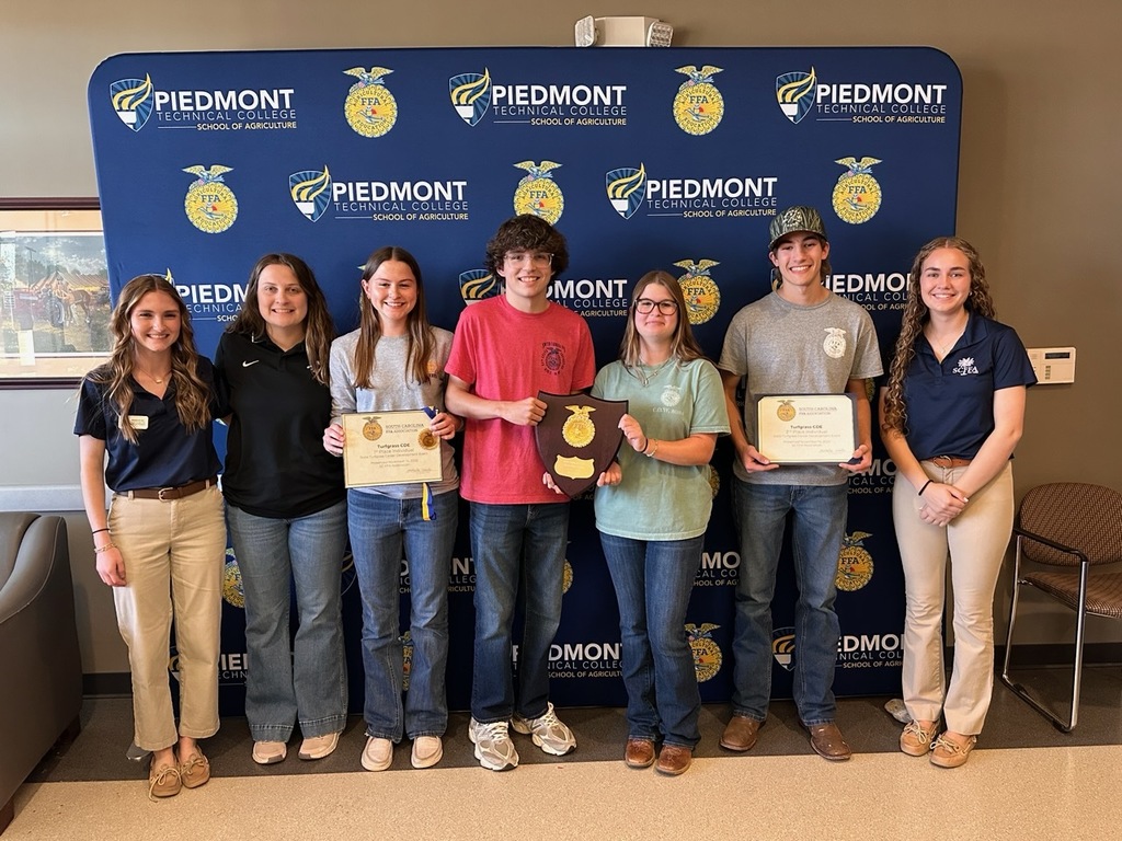 A group of eight young people standing in front of a blue backdrop displaying the Piedmont Technical College School of Agriculture name and FFA emblems. Most are smiling; several hold certificates or plaques. They wear casual clothing (jeans, polos). The scene appears to be an indoor awards or recognition event.