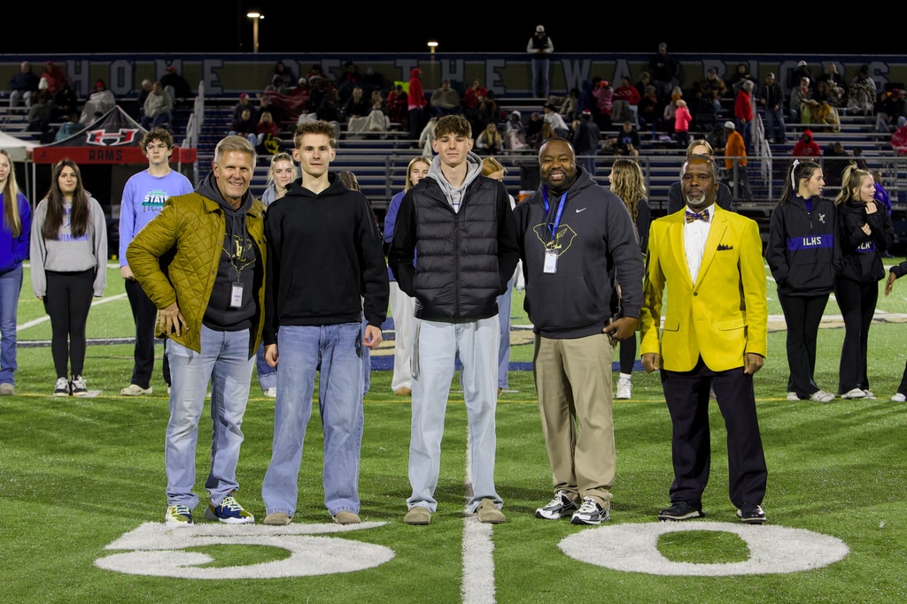 Nighttime photo on a green field near the 50‑yard marker showing five men standing in a row (left to right: a man in an olive quilted jacket; a man in a black hoodie; a man in a black puffer vest over a grey hoodie; a man in a dark grey hoodie and khaki pants; and a man in a bright yellow blazer with a plaid bow tie). Younger people in hoodies and tracksuits stand behind them; bleachers and a wall reading “HOME OF THE RAMS / ILHS” are visible in the background.