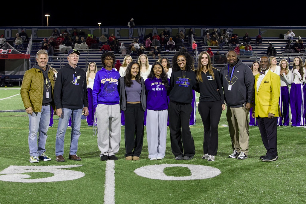  Nighttime field photo near the 50‑yard line with eleven people standing in a row in front of bleachers under stadium lights. A blue wall behind them reads “WELCOME TO THE WARRIORS.” From left to right: an older man in an olive quilted jacket; a man in a black hoodie and cap; a young person in a purple “Spiders” hoodie; a young woman in a grey crop top; a young woman in a purple “INDIAN LAND WARRIORS” hoodie; a young woman wearing a “REGION CHAMPION 2022–2023” hoodie; a young woman in a black shirt with “TENNIS” on the side; a man in a black Nike hoodie with khaki pants; and a man in a bright yellow blazer and bow tie. Red tent labeled “ADAMS” is visible at left; a dance or cheer group in white tops and purple pants stands behind the main group.