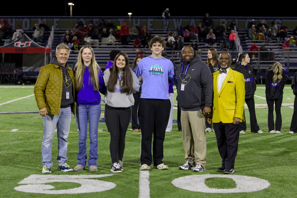 Close group photo showing people wearing light blue t‑shirts that read “2023 SWIMMING STATE CHAMPIONSHIPS ILHS” with other individuals in black jackets marked “ILHS.” Stadium bleachers in the background display a “HOME OF THE WARRIORS” banner.