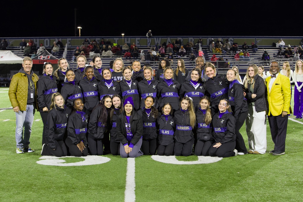 Nighttime team photo on a football field showing 25 people—about 20 young women in black long‑sleeve tops and black track pants arranged kneeling and standing in two rows, with three adult men and two adult women among them. Floodlights illuminate the group; bleachers and a banner reading “HOME OF THE WARRIORS” appear in the background. Several people are smiling and some students wear small facepaint designs.