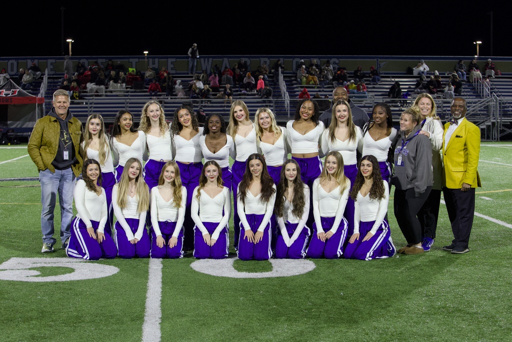 Nighttime team photo on a football field showing 25 people—about 20 young women in white long‑sleeve tops and purple track pants arranged kneeling and standing in two rows, with three adult men and two adult women among them. Floodlights illuminate the group; bleachers and a banner reading “HOME OF THE WARRIORS” appear in the background. Several people are smiling and some students wear small facepaint designs.