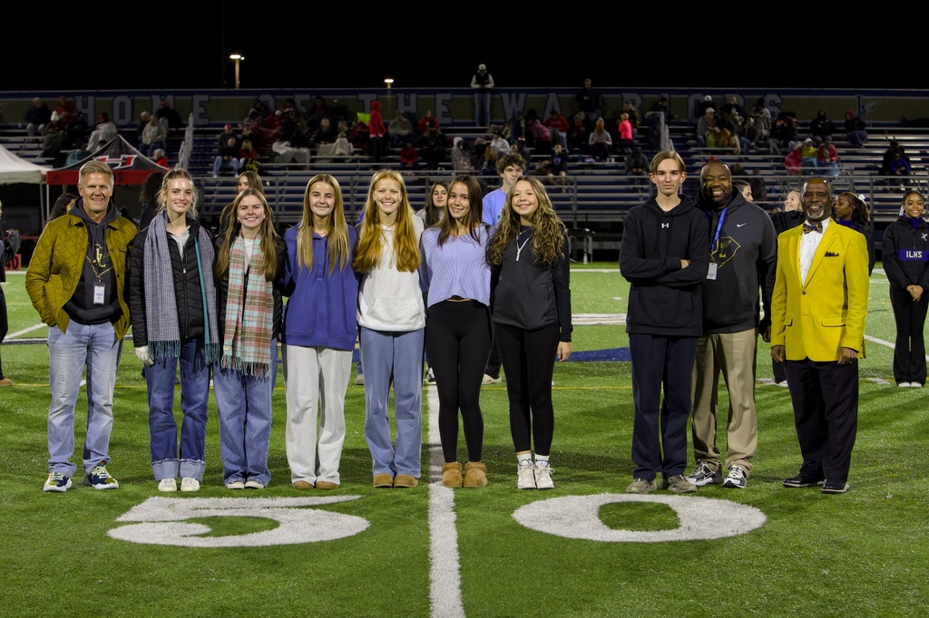 Nighttime photo on a football field near the 50‑yard line showing a line of people standing shoulder to shoulder in front of stadium bleachers and bright lights. From left to right: an adult man in a yellow quilted jacket and jeans; a teenage girl in a dark jacket and blue/white plaid scarf; a teenage girl in a white shirt and plaid scarf; a teenage girl in a blue sweatshirt and light pants; a teenage girl in a white hoodie and light blue pants; a teenage girl in a light blue long sleeve and black leggings; a teenage girl in a black top, black leggings and white sneakers; a teenage boy in a black hoodie with arms crossed; an adult man in a grey hoodie and khaki pants; an adult man in a bright yellow blazer with a patterned bow tie; and a partially visible person wearing a dark top with “ILHS.” Several adults wear lanyards. A large “HOME OF THE WARRIORS / ILHS” sign is visible in the background.