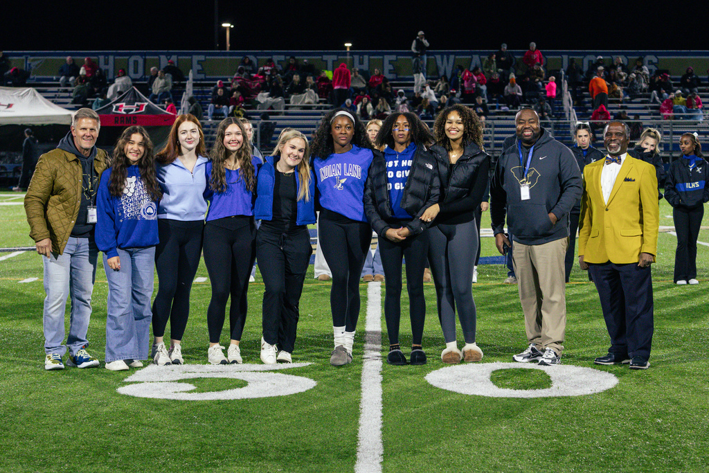  Nighttime group photo on a football field with twelve people lined up along a white field marking and stadium bleachers behind them. The group includes men and young women wearing a mix of jackets and blue sweatshirts—one sweatshirt several read “INDIAN LAND,” and one sleeve shows “ILHS.” A few individuals have face paint. A blue banner and a red banner hang above the stands; the field shows two white circular markings in the foreground.
