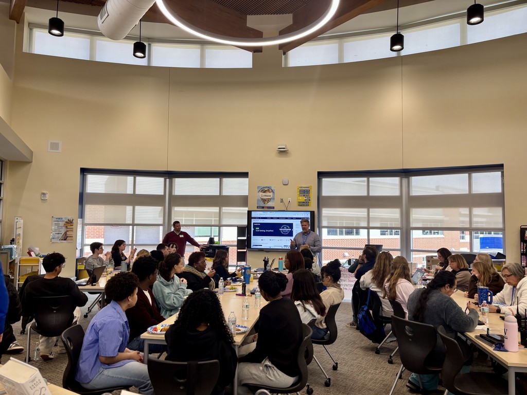 A bright, modern library where students and staff sit at rectangular tables in small groups while two men lead a presentation at the front. A large screen behind them displays the words “Leader In Me.” One presenter in a checkered shirt gestures toward the screen; another stands nearby in a maroon top. Tables hold water bottles and plates. The room has high ceilings with circular and pendant lights, large windows with blinds, light beige walls with posters, and carpeted floors.