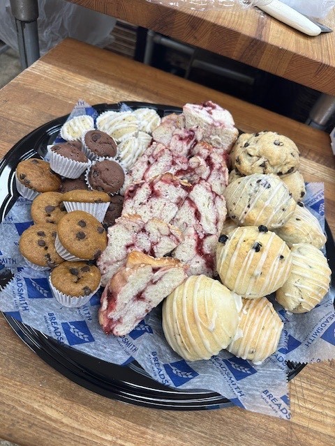 Round black platter filled with assorted baked goods on paper liners printed "BREADSMITH / HAND MADE BREAD." Left side: small chocolate chip cookies and mini chocolate muffins in white liners; light-colored pastries with white icing drizzle. Center: slices of pink/red berry-swirled bread or cake. Right and bottom: various scones or biscuits, some topped or drizzled with white icing and some with chocolate chips.