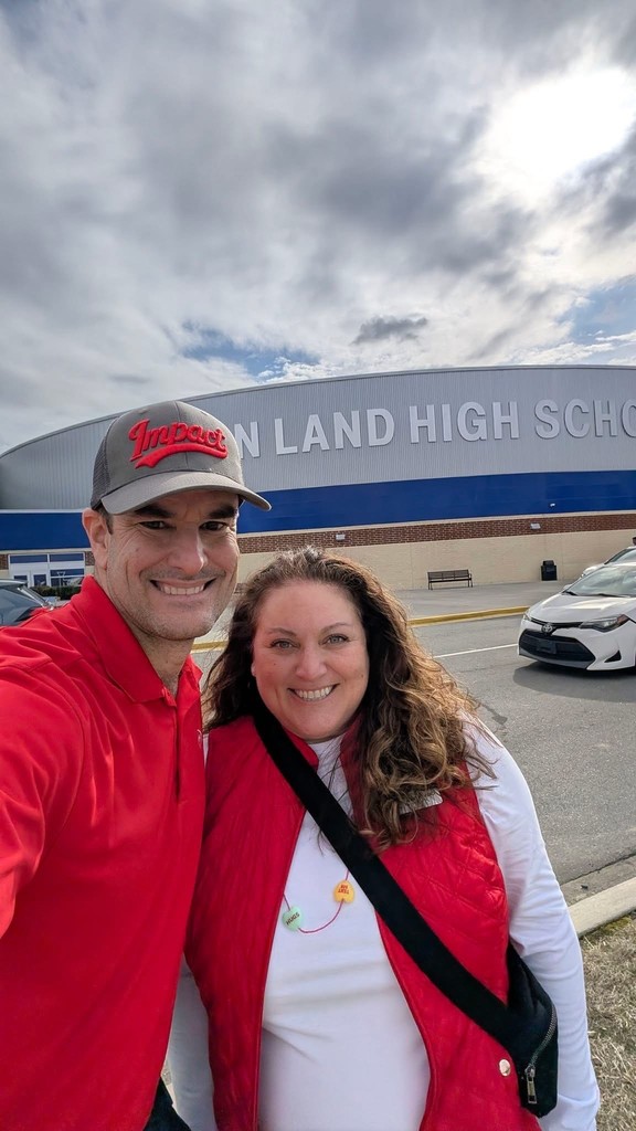 Selfie of a smiling man and woman standing outdoors in front of a large school building. The man (left) wears a red polo and gray cap with "Impact" script; the woman (right) wears a red quilted vest over a white top and a necklace with two heart-shaped charms. Part of the building facade shows "LAND HIGH SCHOOL." Cars are visible in the parking lot and the sky is overcast.