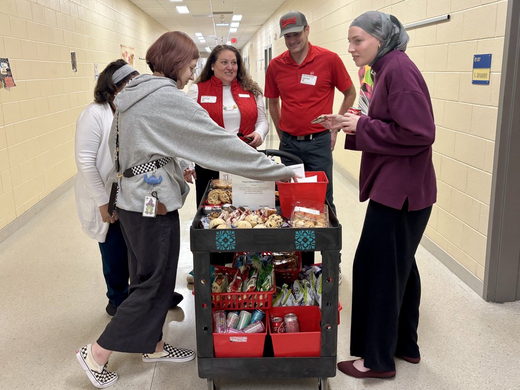 Five people gathered in a bright hallway around a two-tier snack cart. The top shelf holds baked goods; a person with reddish-brown hair in a gray hoodie and checkered Vans reaches toward the treats. To her right are a woman in a red vest and a man in a red polo and cap wearing visitor badges. Another woman in a gray head covering and dark purple top holds a cookie and soda. The bottom shelf has red baskets with chip bags and soda cans. "Please enjoy a treat on us!"