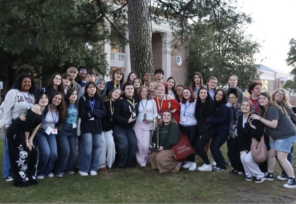 Cast of ILHS Honors Theatre class pose outside of a building on a college campus. They pose with medels and a trophy.