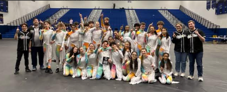 winter percussion poses in a gymnasium on a gray floor, they wear white costumes accented with pastel colors. 
