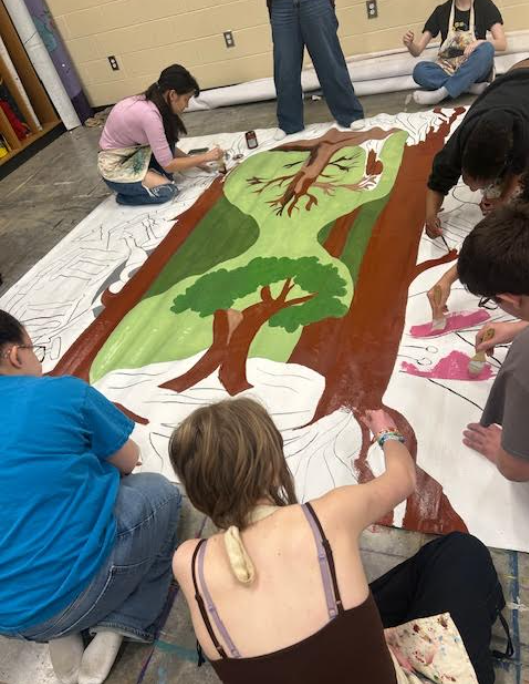 A group of at least six people kneel, sit, and stand around a large mural or banner laid flat on a light floor, collaboratively painting a stylized natural landscape. The design centers on a large brown trunk with branching limbs and broad areas of light and dark green foliage; other sections are being painted pink and many areas remain white with pencil outlines. Paint pots and brushes surround the canvas; the artists wear casual clothes and some wear aprons. The indoor studio background shows a plain light wall, a few small dark electrical outlets, and a rack with rolled materials.