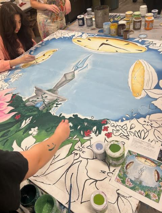 A large collaborative painting spread flat on the floor shows a fantastical landscape: a light blue sky with three elongated golden clocks (appearing to float or melt), a central spiky crystalline formation in grey and blue, and a lush green field with many flowers. Several people paint different sections; some areas (sky, clocks, crystal, parts of foliage) are colored while foreground flowers and leaves remain black‑and‑white sketches. The foreground shows a person’s arm with a green smiley drawn on it painting foliage; another person with long dark hair paints a clock. Many small paint pots and brushes are scattered nearby and a small printed reference image of the finished artwork lies at the lower right.