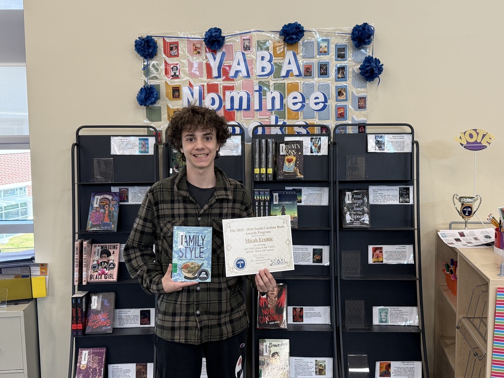 student smiling holding a certificate and book in front of a library shelf. 