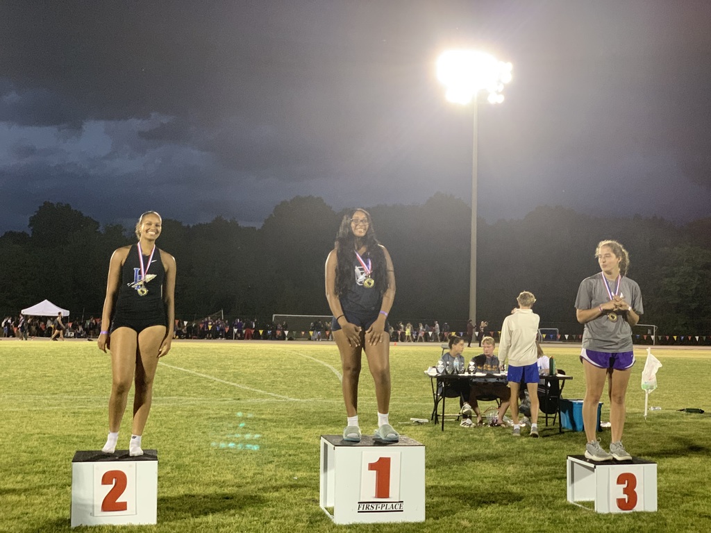  "Three young women on a nighttime awards podium at a track and field event. First-place athlete in center with long dark hair wears dark athletic top and light sandals, gold medal with red, white, and blue ribbon. Second-place athlete on left in black athletic wear with white shoes, gold medal. Third-place athlete on right with curly light brown hair wears gray shirt and purple shorts, gold medal with white and purple ribbon. Spectators and tents visible in background."
