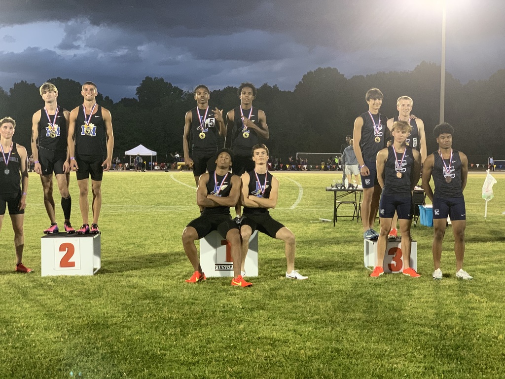  "Male athletes on three-tier awards podium at a nighttime track and field event. First-place podium: four athletes in black singlets with 'LS' logo wear gold medals. Second-place podium: three athletes in black singlets with 'M' logo wear silver medals. Third-place podium: three athletes in dark blue singlets with 'LS' logo wear bronze medals. Stadium lights, white tents, and officials' table visible in background."