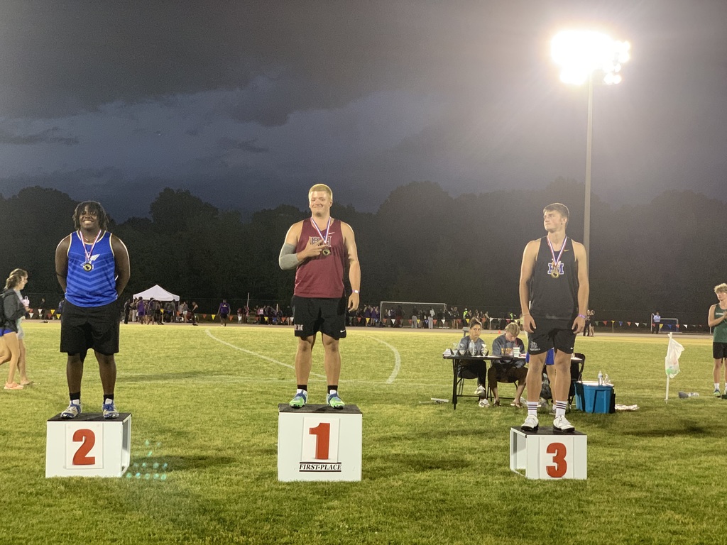 "Three male athletes on a nighttime awards podium at a track and field event. First-place athlete in center wears maroon tank top marked 'IPW' with light grey arm sleeve. Second-place athlete on left wears blue and white striped tank top with winged emblem. Third-place athlete on right wears black tank top. All wear gold medals. Stadium lights and officials' table visible in background."