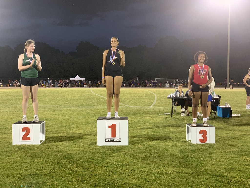 Three female athletes on a nighttime awards podium at a track and field event. First-place athlete in center wears black, holds gold medal, smiling. Second-place athlete on left in green tank top claps. Third-place athlete on right in red tank top wears gold medal. Stadium lights and spectators visible in background."