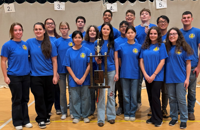 Seventeen ILHS JROTC team members in matching royal blue t-shirts with yellow and blue logos pose on a gymnasium floor. A young woman in the center holds a tall silver trophy on a dark wooden base. Numbered white cards '2,' '3,' '4,' and '5' hang on the light-colored wall behind the group."