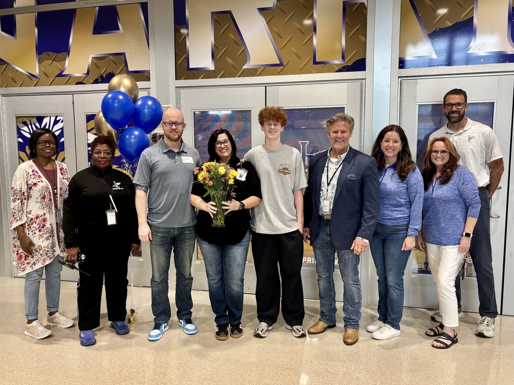 Group of ten individuals (six women and four men) standing in a line, smiling and posing for a photograph indoors in front of a wall with large decorative letters in gold and blue partially spelling "START." Blue and gold balloons visible on the left side. Woman, Ms Alm,  in the center holds a clear glass vase with a bouquet of yellow and orange flowers. All individuals dressed in a mix of casual and business-casual attire in what appears to be an event or school hall setting.