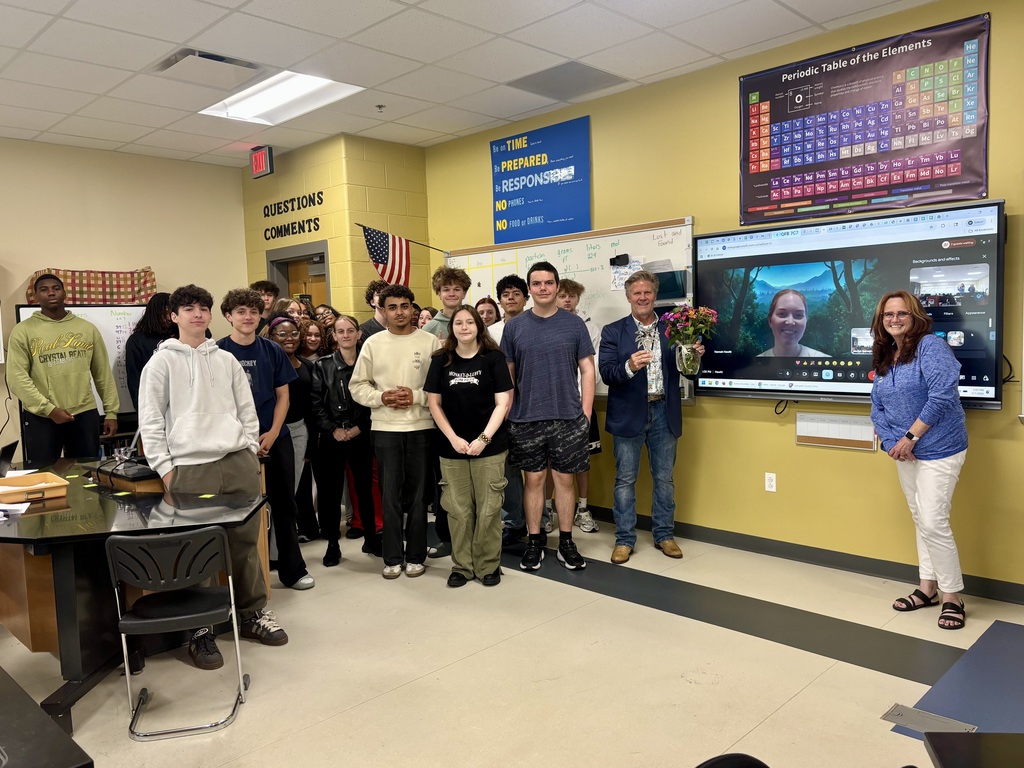 Classroom scene with yellow walls showing approximately 20-25 students and two adults celebrating an award. Large digital screen on right wall displays a video call with a smiling young woman against a forest background. Man in blue blazer stands on right holding clear trophy or vase filled with colorful flowers, with woman in blue top and white pants standing beside him smiling at camera. American flag hangs on wall. Whiteboard with handwritten notes visible showing "Crystal React, grams, moles, g/mol" and percentages. Classroom rules poster displays "Be on TIME, Be PREPARED, Be RESPONSIBLE, NO PHONES, NO FOOD or DRINKS" with "QUESTIONS COMMENTS" at top. Large Periodic Table of the Elements poster prominent on upper right wall. Desk and black chair in foreground on left side. Light-colored floor with dark grey stripe and white ceiling with fluorescent lighting visible.