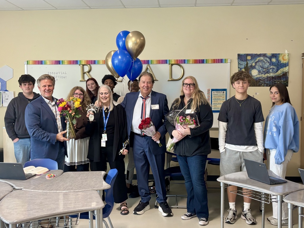 Group of approximately eleven people including adults and students gathered in classroom for celebratory award event. Background features whiteboard with rainbow border and large golden letters spelling "RAD." Van Gogh's "The Starry Night" print visible on far right wall. "ESSENTIAL QUESTION:?" and "WARRIOR UP &" text visible on wall. From left: young man in black hoodie stands next to adult man in blue suit jacket holding vase with colorful bouquet; woman with long blonde hair and glasses; woman with white hair in black top holding small silver pineapple-shaped trophy; adult man in blue suit with red, white, and blue striped tie and name tag holding bouquet of red roses; woman with long blonde hair and glasses in black jacket holding bouquet of pink and purple flowers. Several students visible in background including young woman with red hair. Far right shows young man in dark t-shirt over long-sleeved white shirt with grey shorts and young woman in light blue sweatshirt with "S.S. ELS" text. Student desks and chairs in foreground with open laptops visible, one desk has book and colorful candy next to laptop.