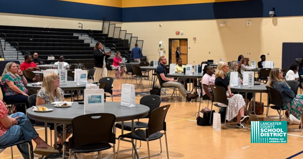 A large indoor room with tables and chairs. People sit around the tables and some stand.