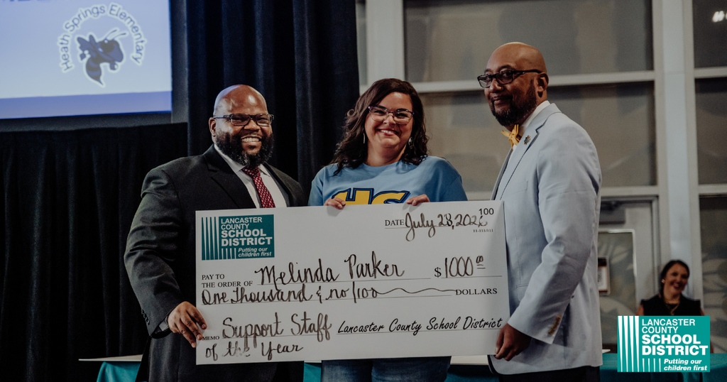 Three individuals on stage holding a large check, one woman smiling, with a backdrop showing logos.