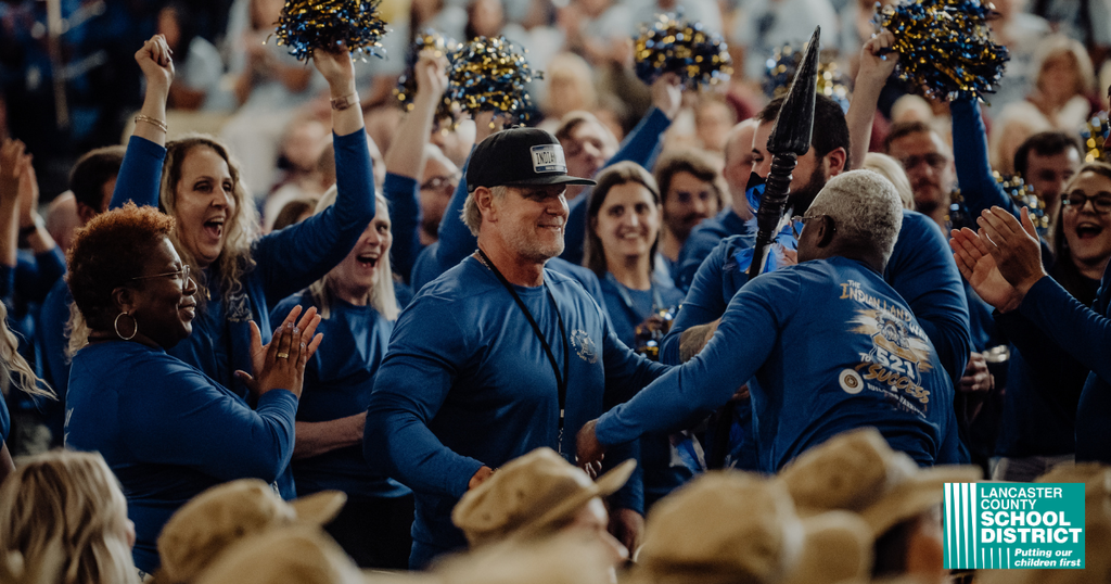 A crowd of people in blue tops and hats, some holding pom-poms, cheer in an outdoor stadium.