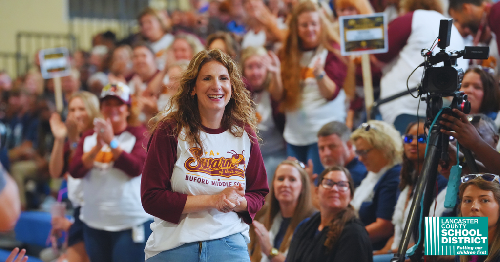 A woman with a white shirt and maroon sleeves stands smiling in front of a cheering crowd.