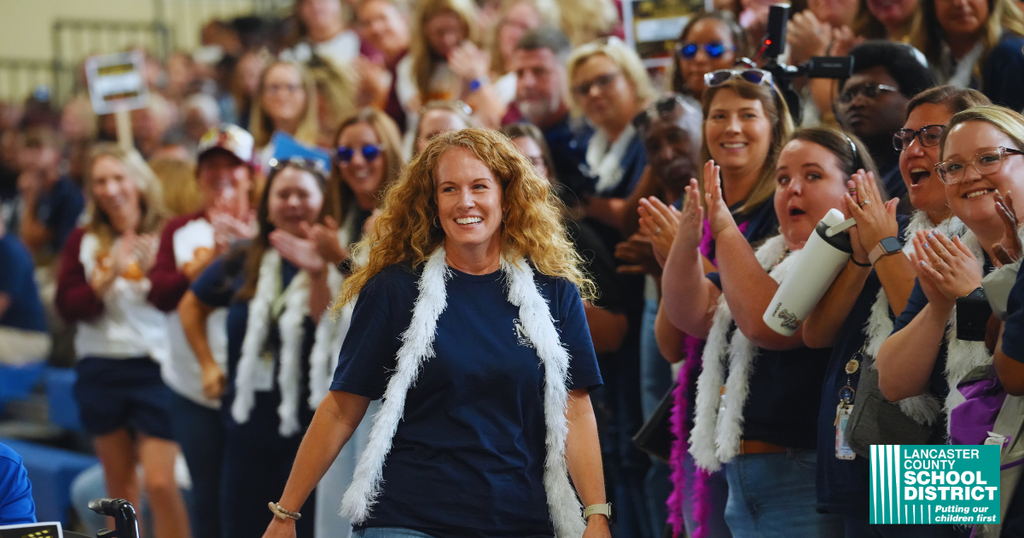 A crowd of people in stands, cheering and clapping. A woman in a blue shirt walks in front.