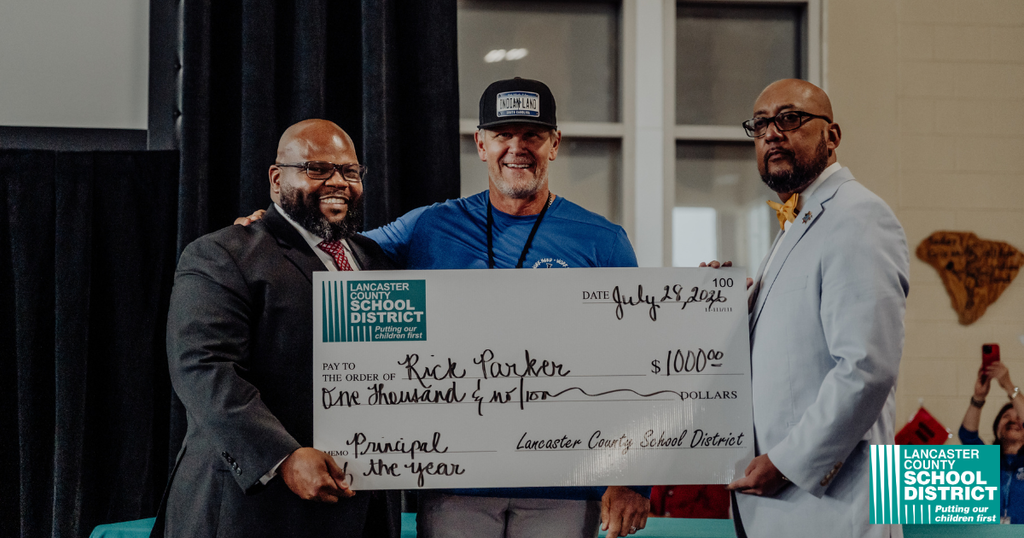 Three men in suits hold a check for $1,000. The man in the middle wears a cap.