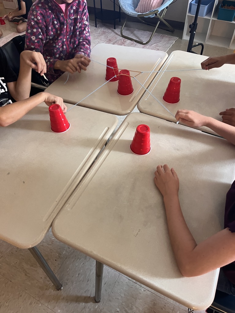 Students sitting around tables playing a game with red cups and string. White tables and carpeted floor.