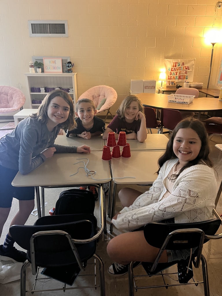 Four girls sit at tables in a classroom, smiling at the camera, with cups in front of them.