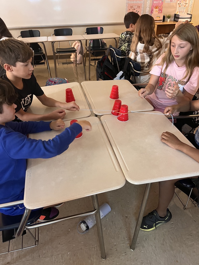 Children in a classroom, seated at desks, engaging in a game using red plastic cups, in a brightly lit room.