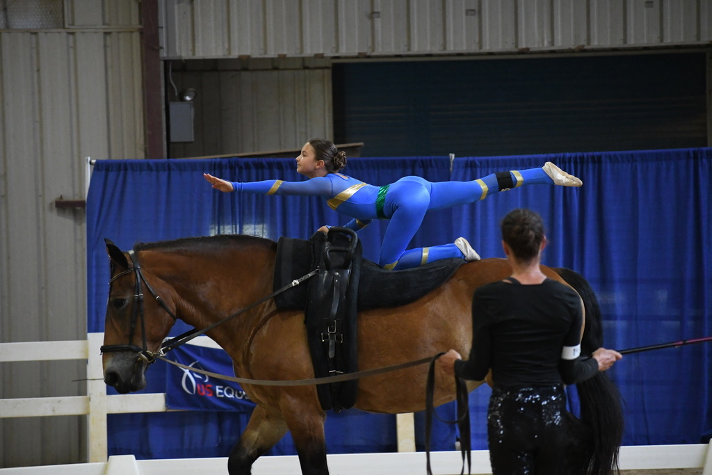A woman performs an acrobatic move atop a horse. She wears blue and gold clothing. Behind her, another person stands.