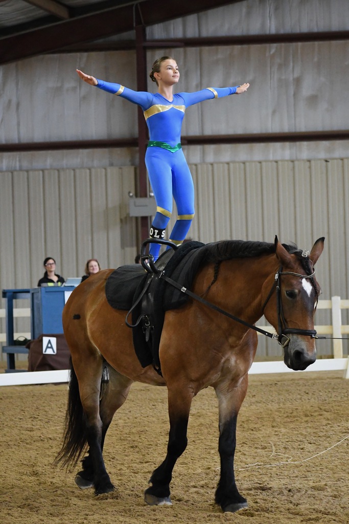 A woman stands on a horse while balancing her arms and legs. She wears a blue jumpsuit.