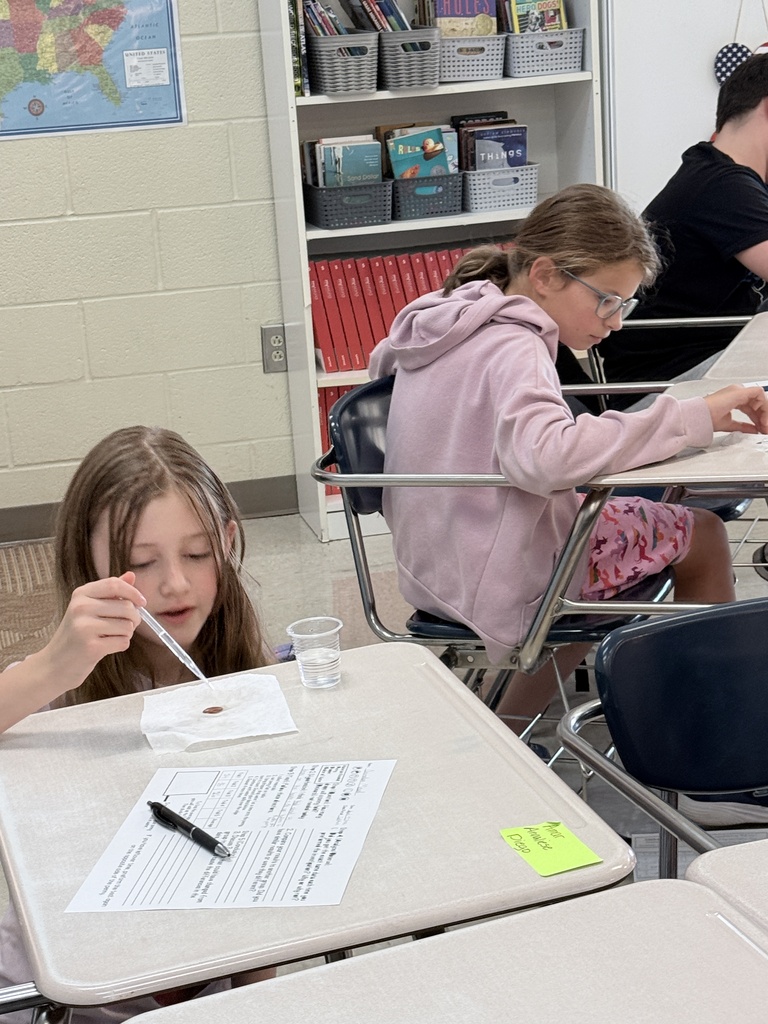 A classroom with desks, one student holding a pipette, another with glasses, and a shelf with books.