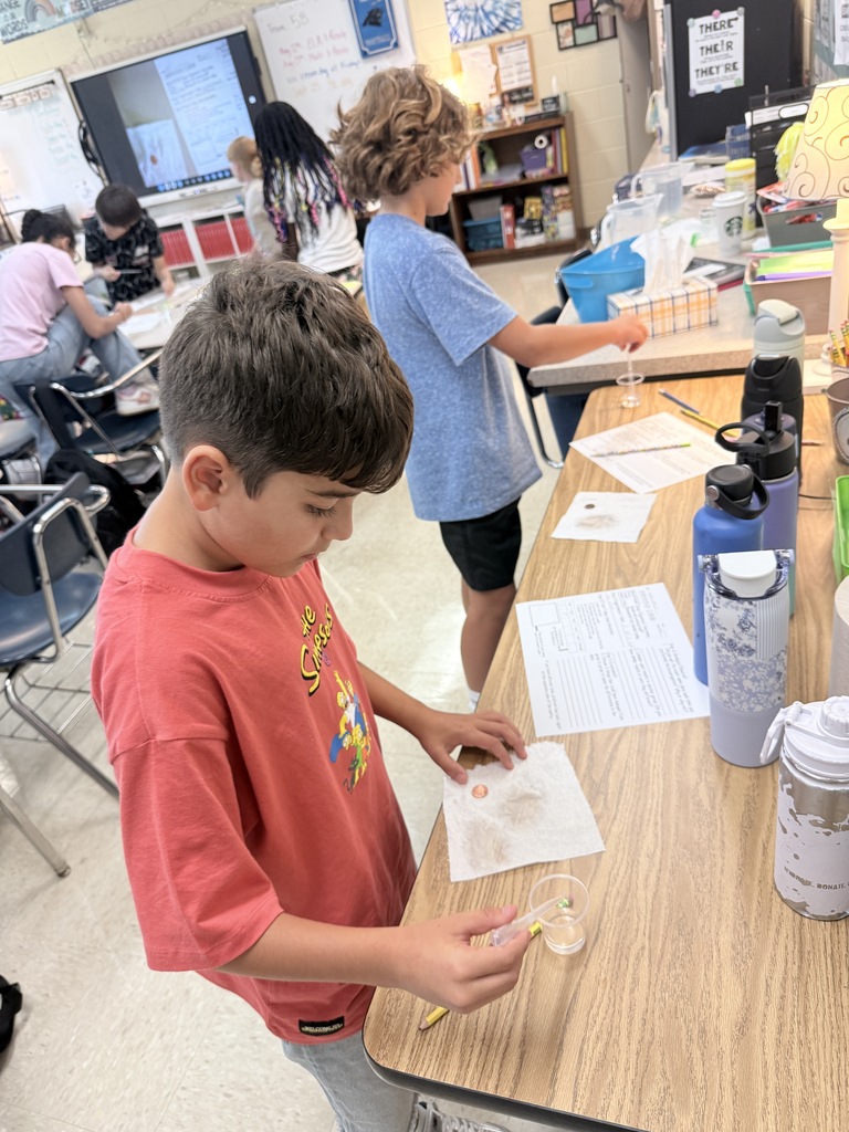 Kids are in a classroom. One boy is holding a container with liquid. Another boy is pouring a liquid.
