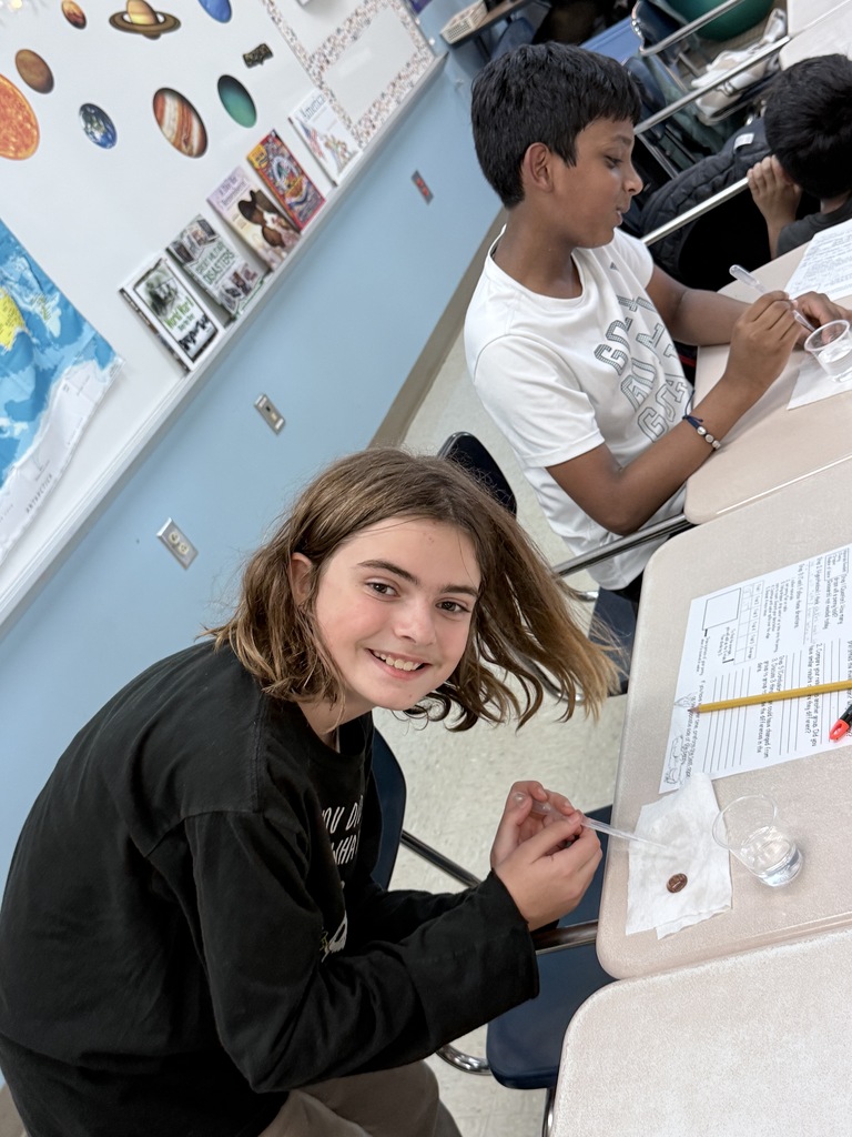 Three children in a classroom, seated at desks. One girl leans over her desk, smiling, and using a pen.