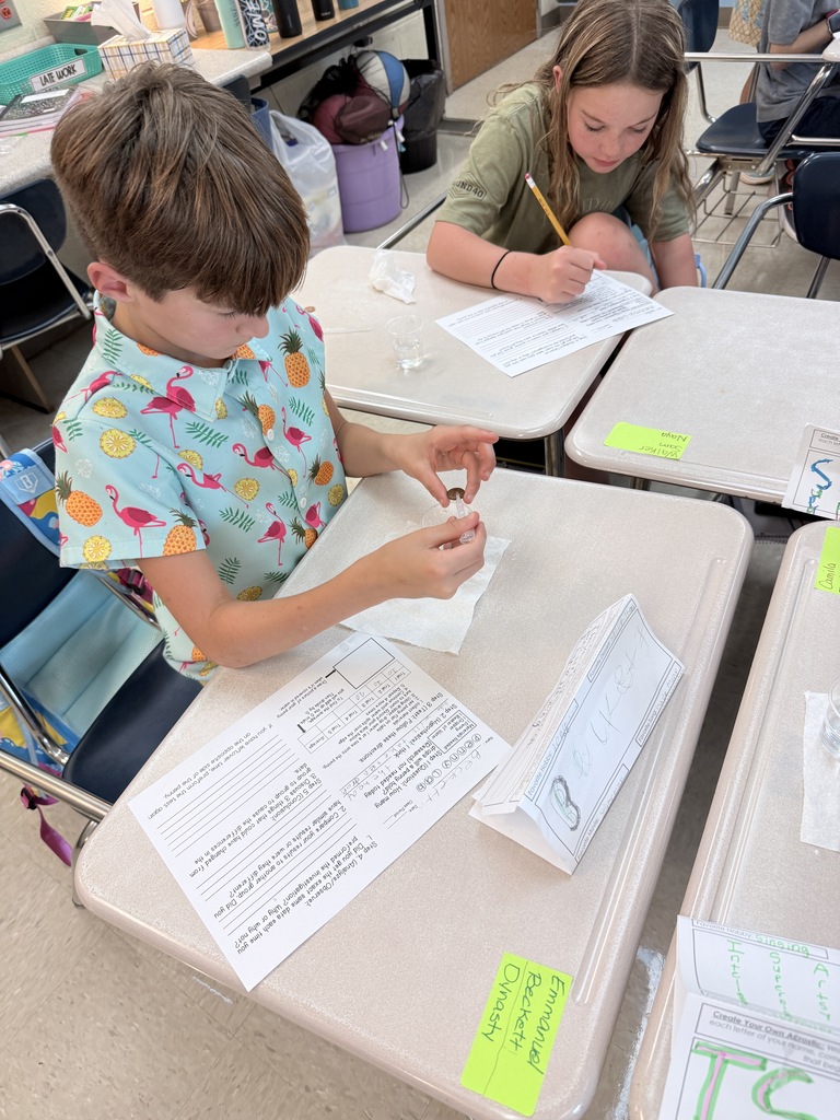 Two children seated at desks in a classroom, one holding a pencil and writing, the other using a pipette. Papers and books on the desks.