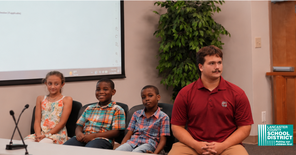 Three children and a man sit at a table in front of a projector screen and plant.