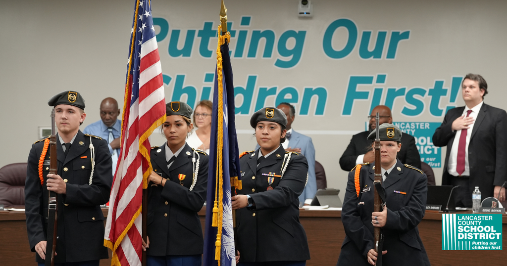 Four cadets hold flags in a room. A sign reads, "Putting Our Children First." A man in a suit stands behind them.