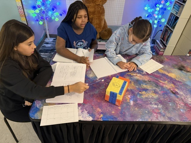 Three students seated at a table with papers and a small box on the table.