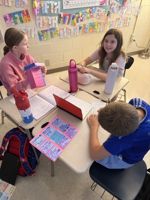 Three children sit at desks in a classroom, with colorful papers on walls behind them.