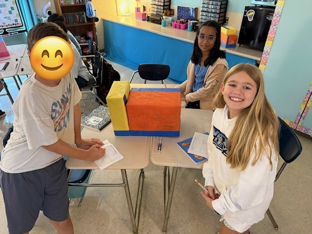 A boy and girl are seated at a table, smiling. The boy wears a face mask with a smiley face.