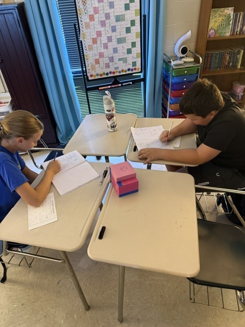 Two students sit at desks in a classroom, each with papers and pens, engaged in writing.