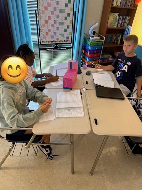 Three children seated at desks in a classroom. One has a smiley face sticker on their head.