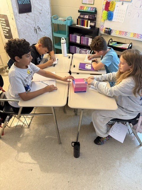 Four children in a classroom sit at desks, working on paper and interacting. Desks are white.